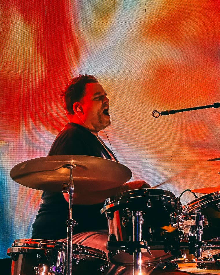 Mario playing drums on stage with The Pollies, mid-hit, in front of an orange and red LED wall with a blurred cymbal in the foreground