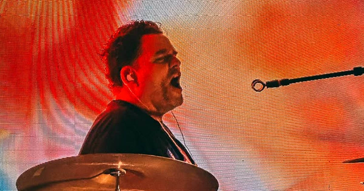 Mario playing drums on stage with The Pollies, mid-hit, in front of an orange and red LED wall with a blurred cymbal in the foreground
