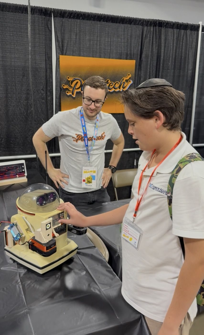 A kid reaching out to touch Ring-O's domed eye on the booth table at Maker Faire Miami while PLACITECH looks on, a PLACITECH banner behind them